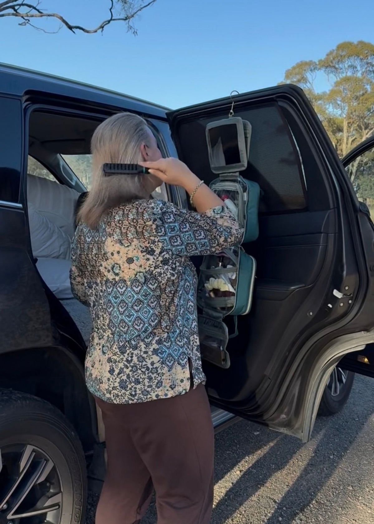 Woman brushing her hair on a road trip using the GLASARI On-The-Glow Cosmetic Case in Ocean Emerald hung from the car door, mirror out