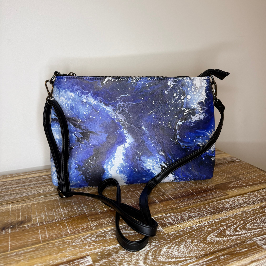 GLASARI Art Cross-Body Bag in Ocean Swirl, a deep blue and white marbled artwork, with its black shoulder strap on a timber table