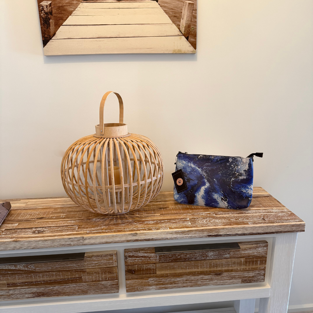 GLASARI Art Cross-Body Bag in Ocean Swirl, a deep blue and white marbled artwork, on a timber side table beside a woven lantern