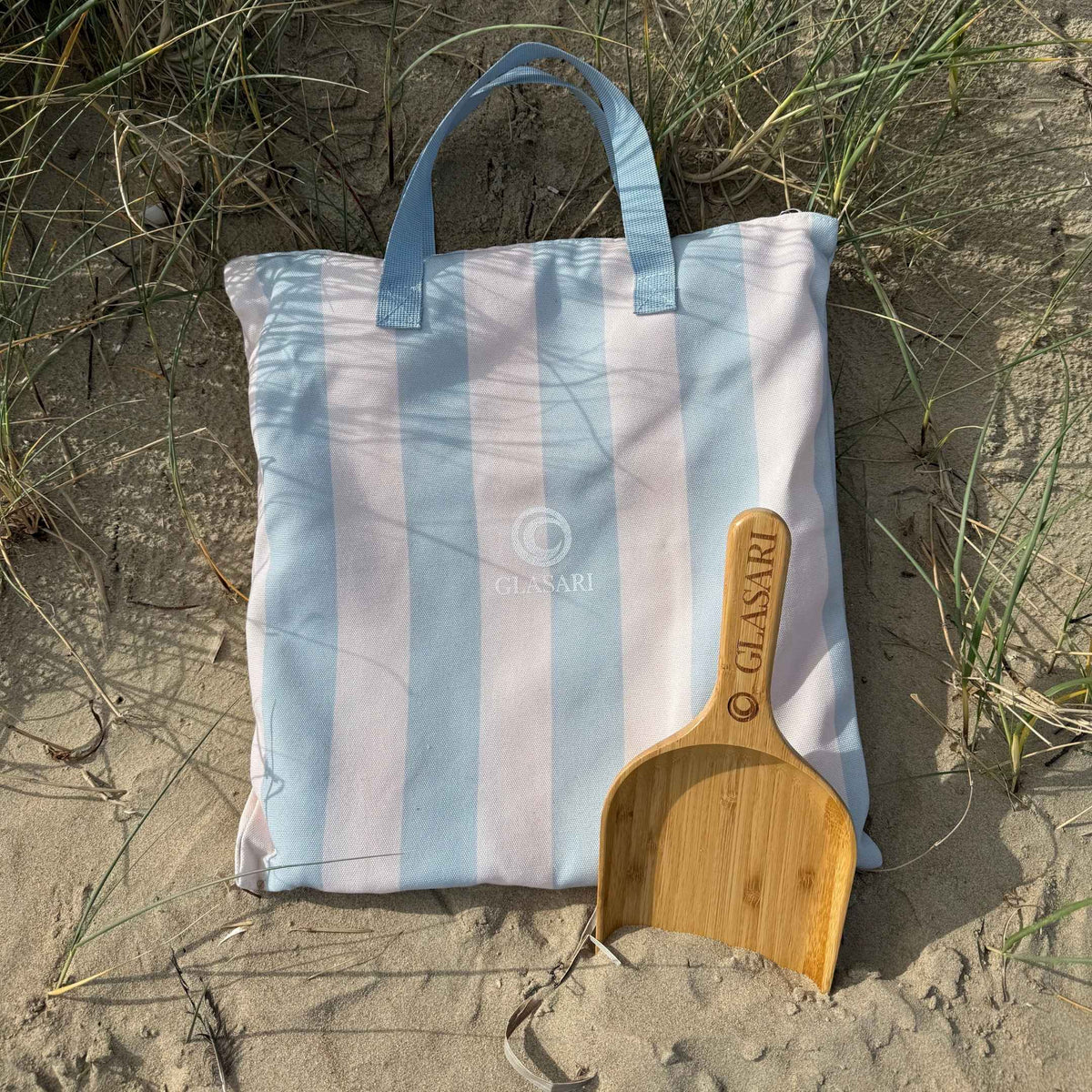 GLASARI Beach Buddy Vault matching carry bag in Blue Horizon Stripe with brown webbing handles, lying in the sand dunes with the free bamboo scoop