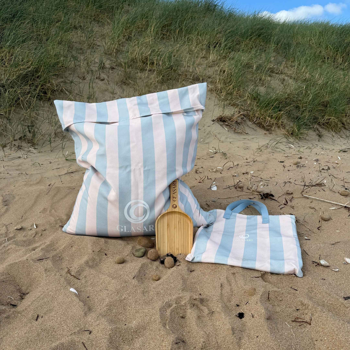 GLASARI Beach Buddy Vault in Blue Horizon stripe filled with sand, with its matching zip carry bag with webbing handles and the free bamboo scoop, in the sand dunes
