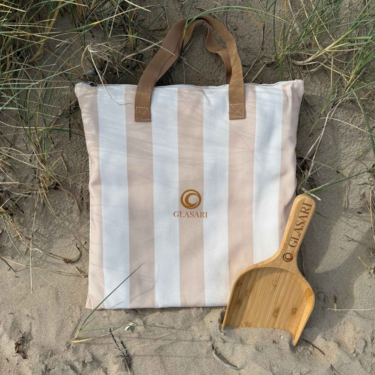 GLASARI Beach Buddy Vault matching carry bag in Coastal Sand stripe with brown webbing handles, lying in the sand dunes with the free bamboo scoop