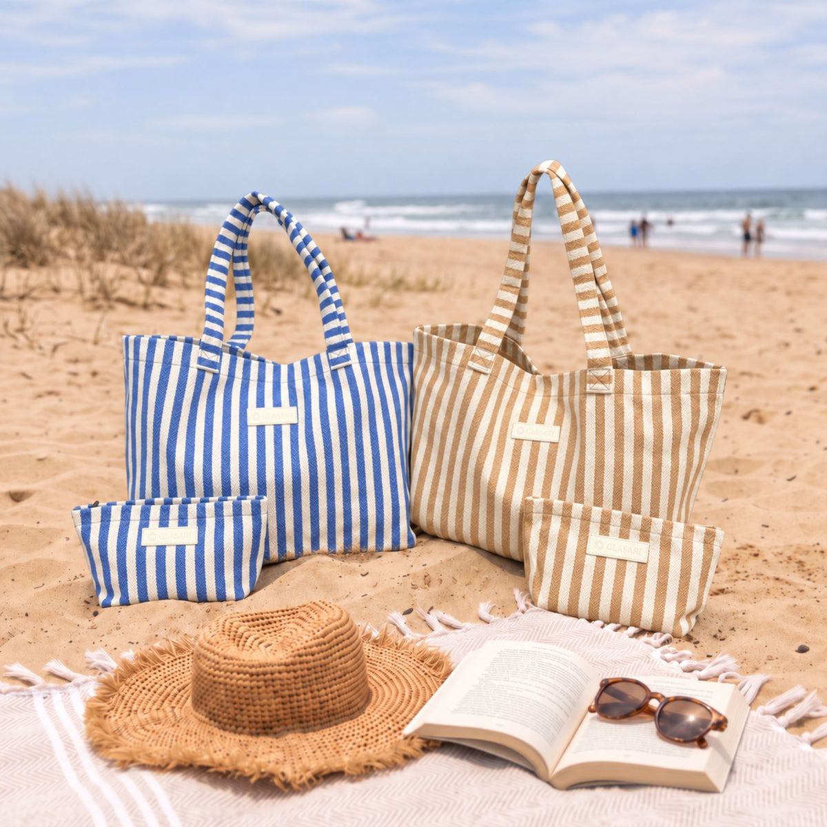 GLASARI Shore Tote in Ocean Blue Stripe and Sand Dune Stripe with matching pouches on a cream beach rug with a straw hat, book and sunglasses