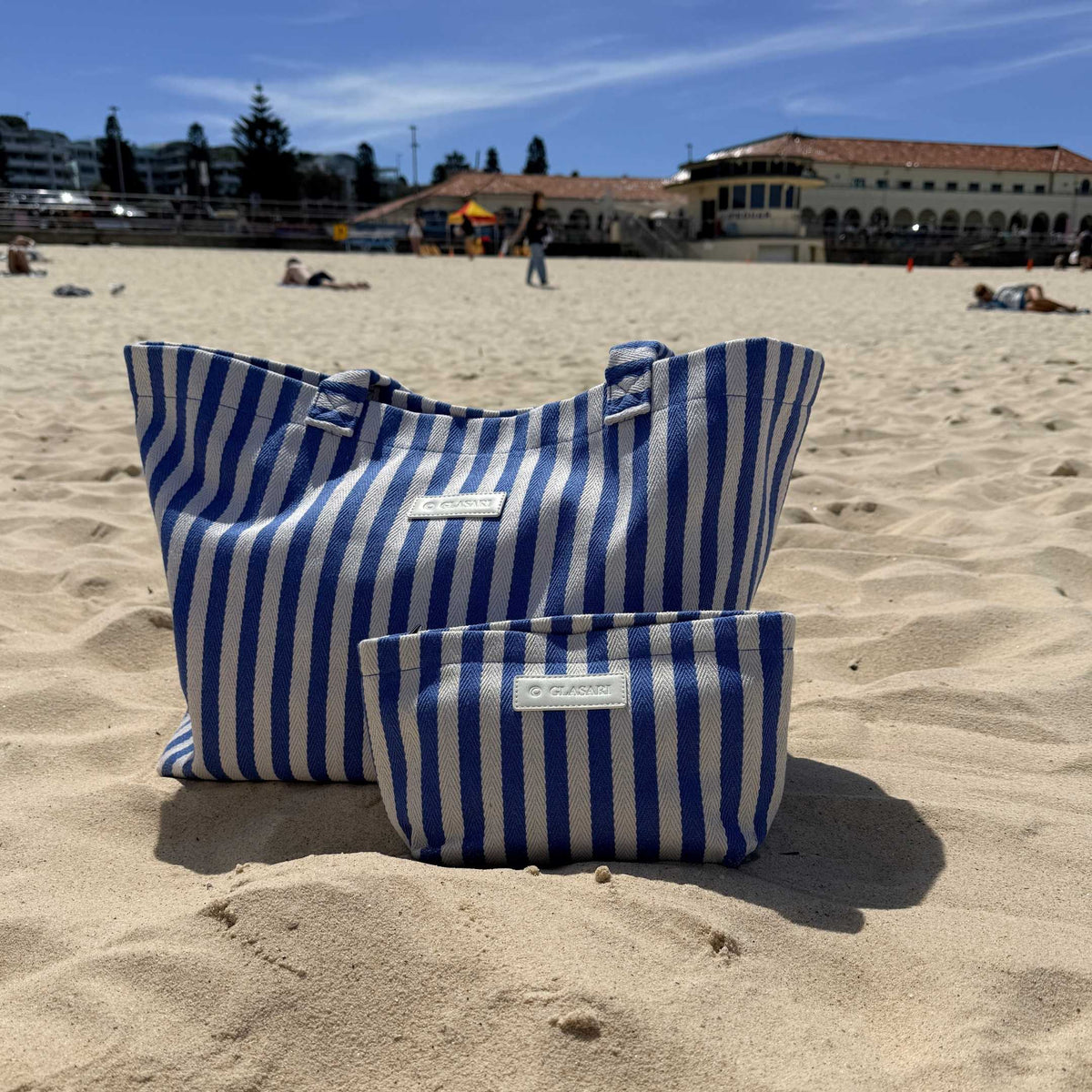 GLASARI Shore Tote and matching pouch in Ocean Blue Stripe sitting on the sand at Bondi Beach, Sydney, with the Bondi Pavilion behind