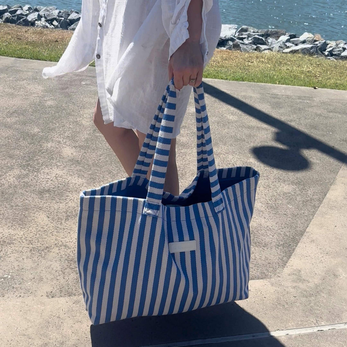 Woman in a white linen shirt carrying the GLASARI Shore Tote in Ocean Blue Stripe by its handles along a sunny waterfront path