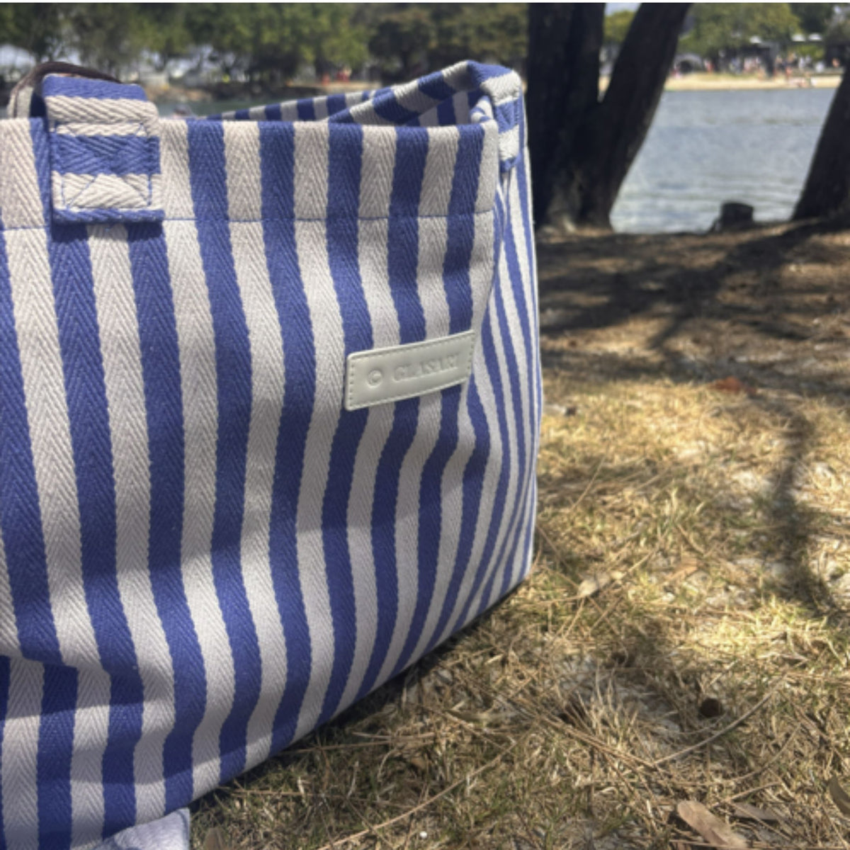 Close-up of the GLASARI Shore Tote in Ocean Blue Stripe resting on the grass by the water, showing the herringbone cotton weave and logo patch