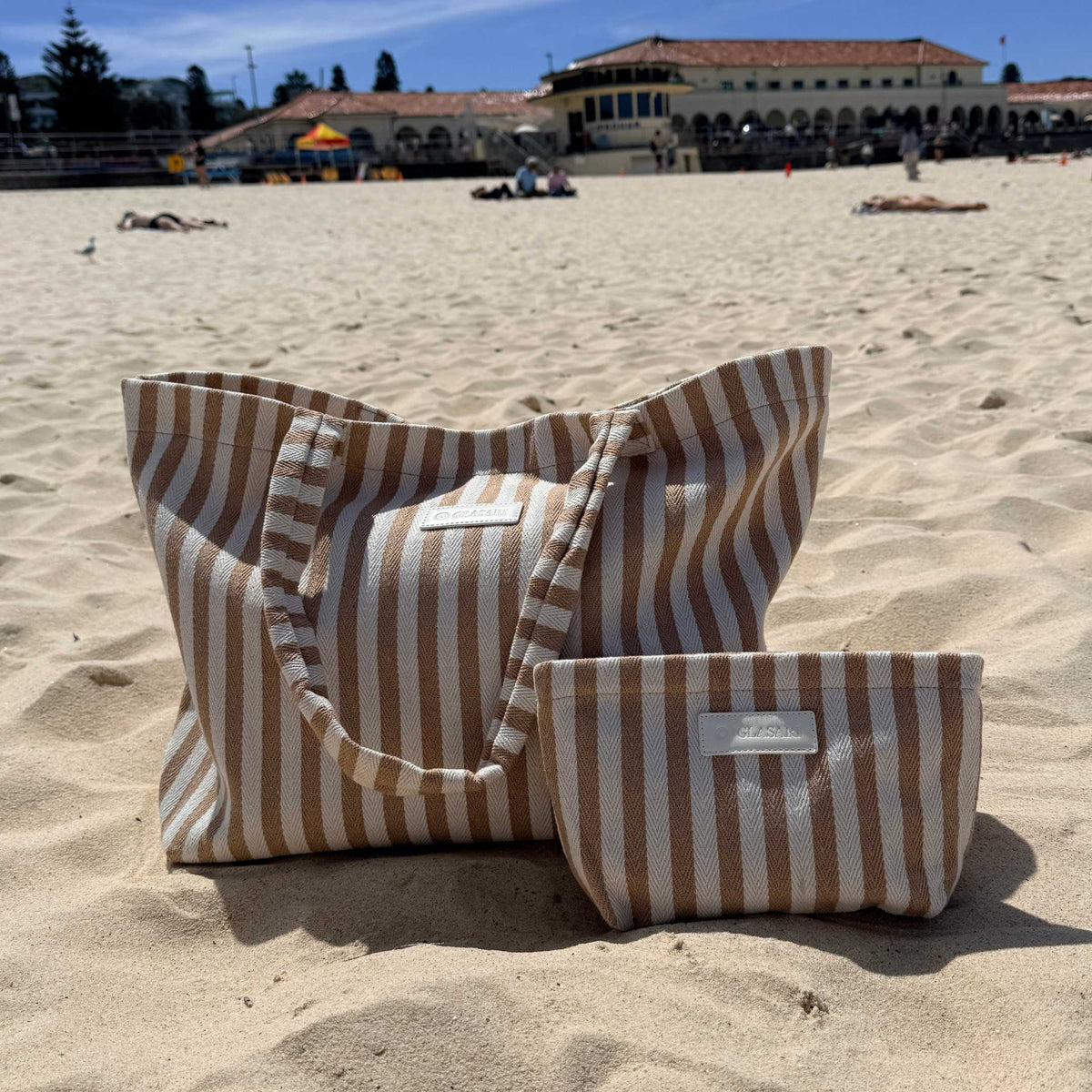 GLASARI Shore Tote and matching pouch in Sand Dune Stripe sitting on the sand at Bondi Beach, Sydney, with the Bondi Pavilion behind
