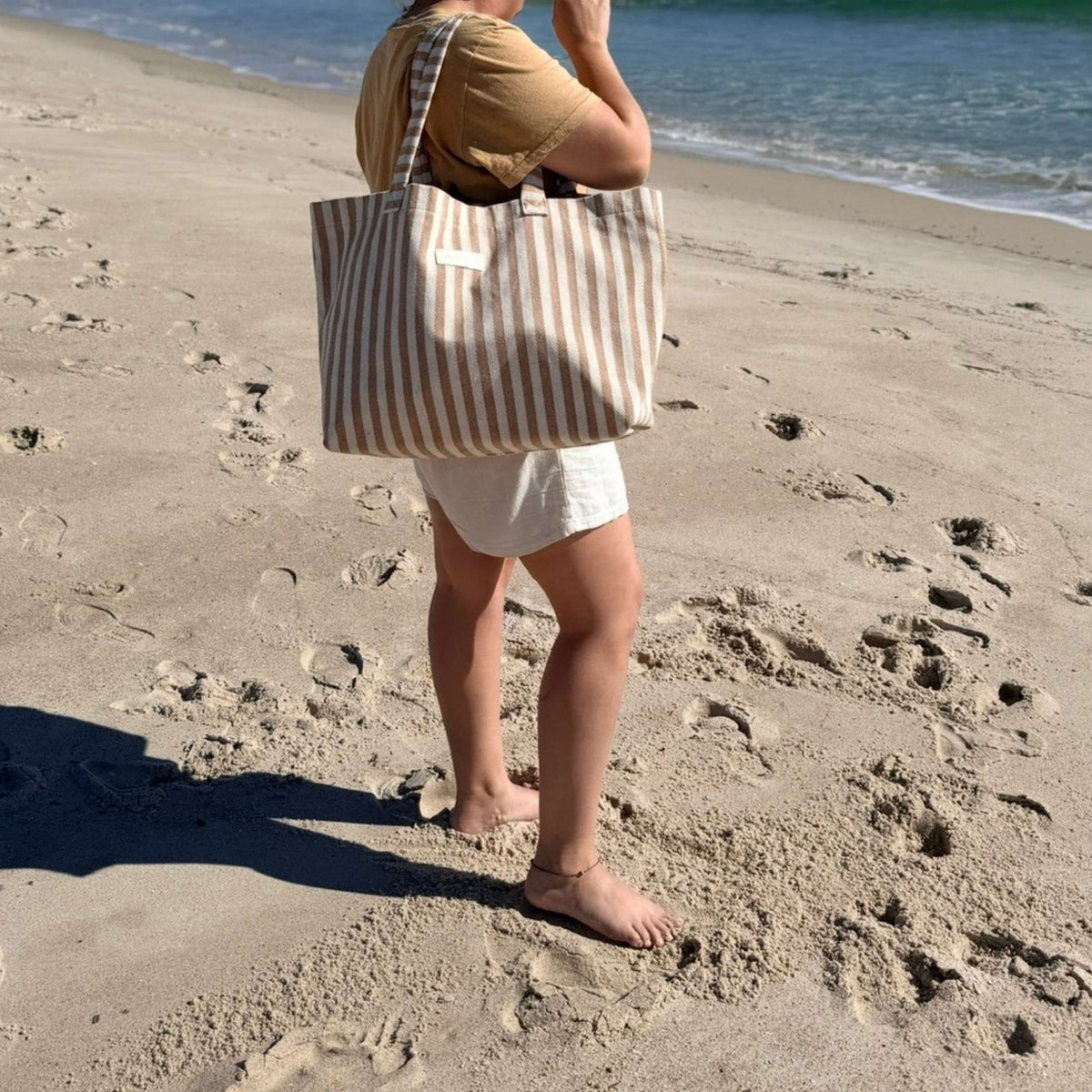 Woman walking barefoot on the beach carrying the GLASARI Shore Tote in Sand Dune Stripe over her shoulder, showing its roomy size