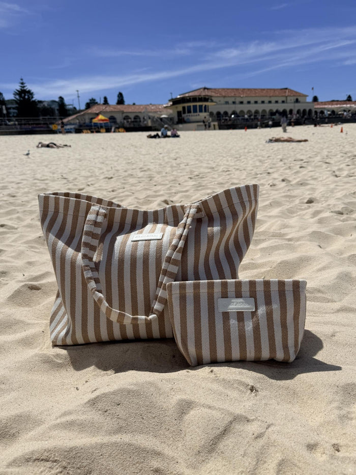 The Shore Tote in sand stripe on the sand at Bondi Beach with the Bondi Pavilion behind